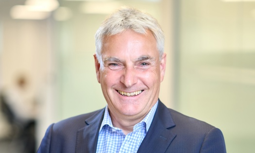 Head and shoulders photograph of a Bromford board member in an office wearing a blue suit and chequered blue and white shirt