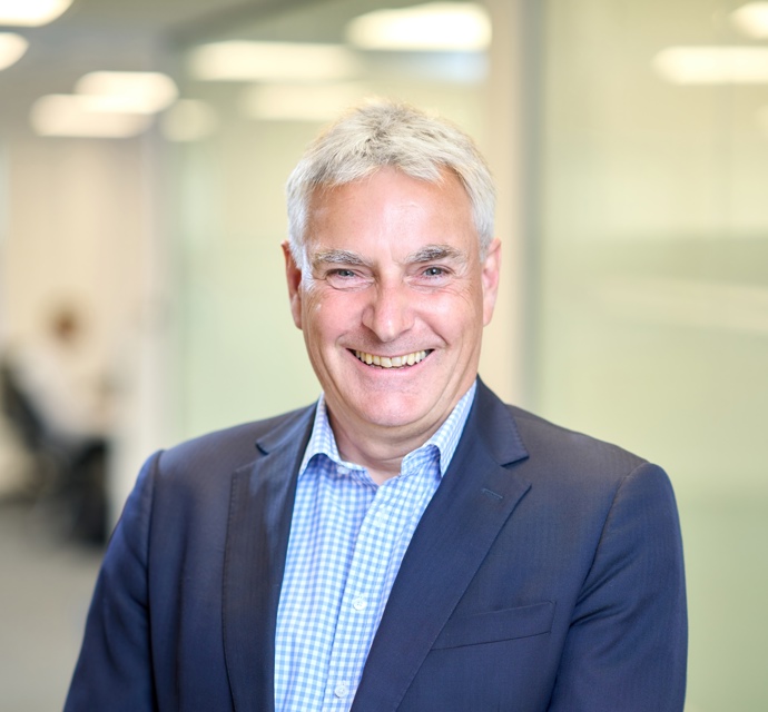 Head and shoulders photograph of a Bromford board member in an office wearing a blue suit and chequered blue and white shirt