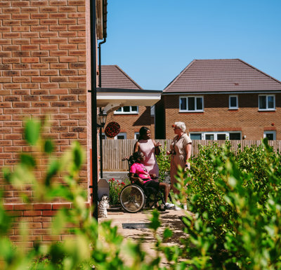 3 people outside a newly built home, two people are standing and a third is sitting in a wheelchair