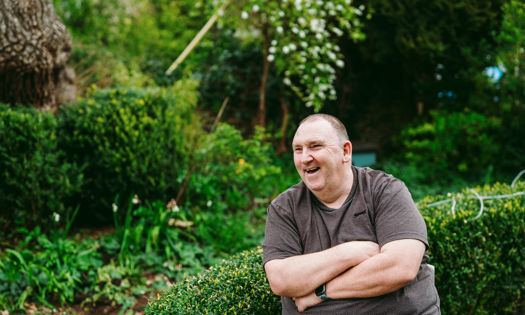 A man sits outside with a t-shirt in a garden, the garden has spring flowers in the background