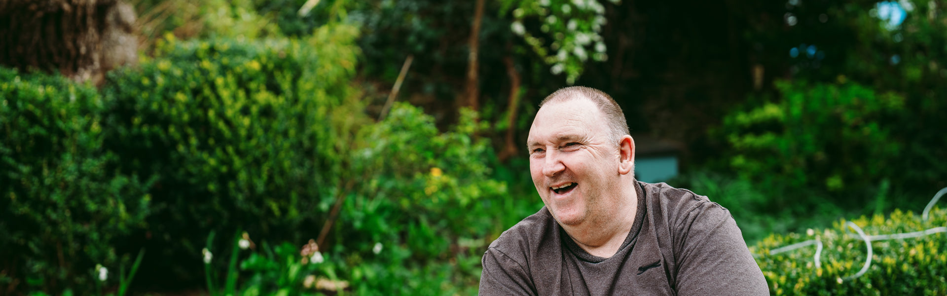 A man sits outside with a t-shirt in a garden, the garden has spring flowers in the background