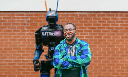 A man stands with arms crossed in front of a robot model