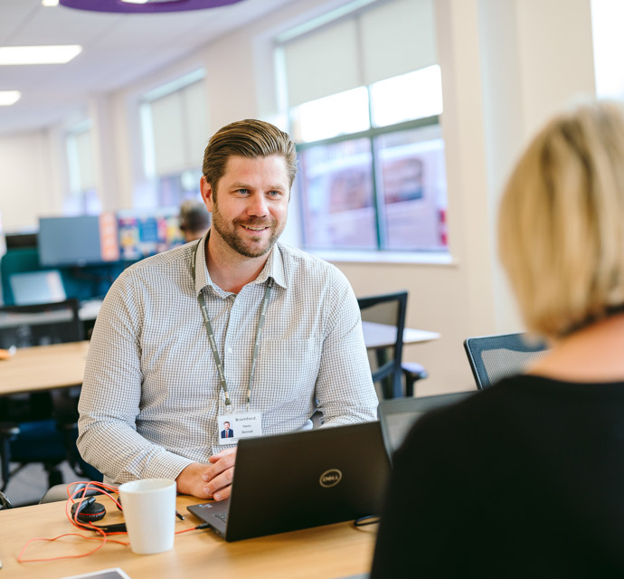 Two Bromford colleagues in an office talking across a desk while working on their laptops