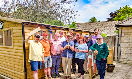 A group of 12 people stand in front of a shed