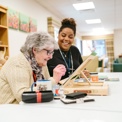 An older lady adds detail to her painting while a younger smiling woman looks on